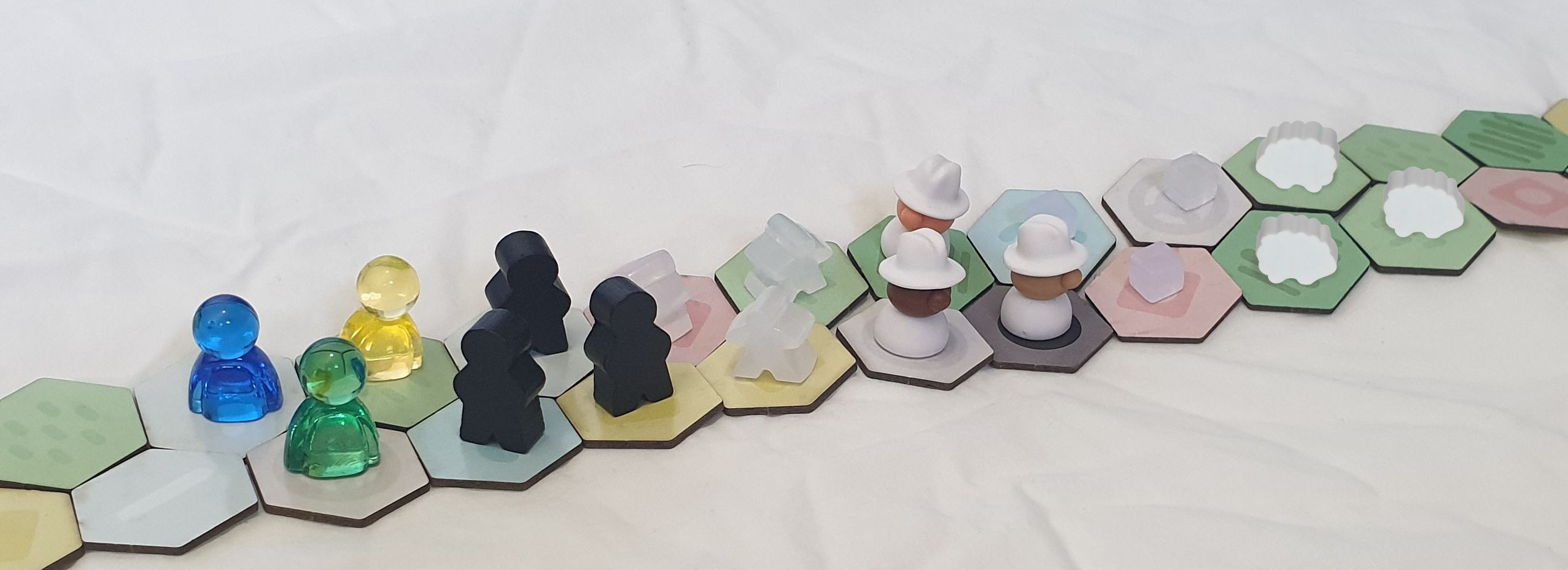 The playing figures laid out over a rolling strip of land tiles. Translucent colorful slimeguys, tall black meeples, squat meeples that appear to be made of pearlescent glue stick, three little men dressed all in white, three cubes, and three wooden sheep